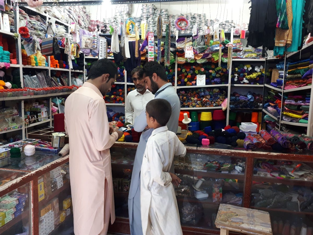 داخل مغازهٔ پارچه و خرازی در بازار چابهار؛ مردان با لباس بلوچی و یک پسربچه