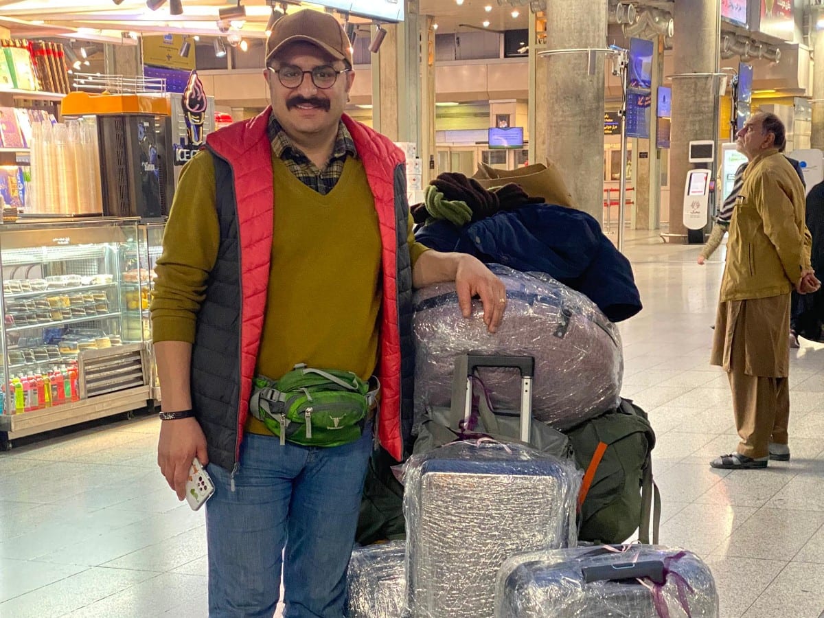 بابامحمد در فرودگاه با چرخ‌دستی پر از چمدان‌های سلفون‌پیچ‌شده